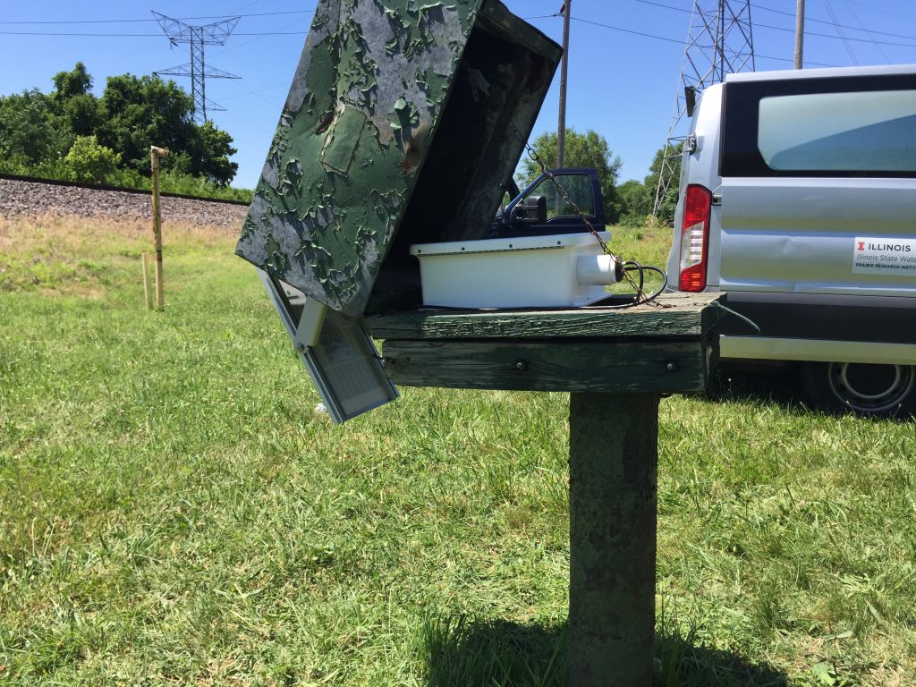 A telemetry monitoring setup with the box lid open. A solar panel hangs off the back of the protective casing, while inside resides a control box of electrical equipment. ISWS field vehicles are in the background.