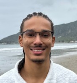 Portrait of Aidan Andrews smiling outdoors at a beach. He has braided hair pulled back, wears glasses, and is dressed in a light-colored sweater. The ocean shoreline and distant hills are visible in the background.