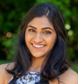 Portrait of Drshika Asher smiling outdoors. She has shoulder-length dark hair and is wearing a sleeveless patterned top. The background is softly blurred greenery, creating a bright, natural setting.