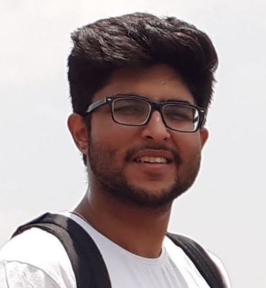 Portrait of Hetarth Chopra smiling outdoors. He has short dark hair, a trimmed beard, and is wearing glasses, a light-colored T-shirt, and a backpack. The background is bright and softly blurred, suggesting an outdoor setting.