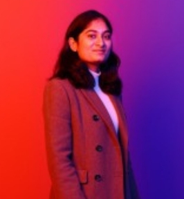 Portrait of Asmita Dabholkar standing and smiling slightly at the camera. She has shoulder-length dark hair and is wearing a brown coat over a light-colored top. The background features a colorful gradient of red and purple lighting, giving the image a modern, studio-style appearance.