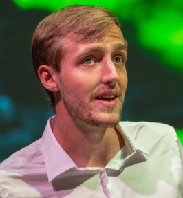 Portrait of Kastan Day speaking or mid-expression at an event. He has short light brown hair and light facial hair, and is wearing a white button-down shirt. The background features green stage lighting, suggesting a conference or presentation setting.