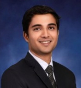 Portrait of Vardhan Dongre smiling at the camera. He has short dark hair and is wearing a dark suit jacket, white dress shirt, and striped tie. The background is a blue studio-style backdrop, giving the image a formal, professional headshot appearance.