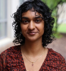 Portrait of Aruna Gauba looking at the camera. She has short, curly dark hair and is wearing a patterned rust-colored top and a delicate necklace. The background is softly blurred greenery and a light-colored wall, suggesting an outdoor or courtyard setting.