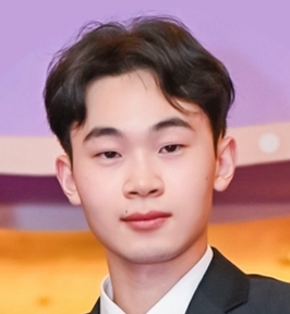 Portrait of Chi Gui looking at the camera. He has short dark hair and is wearing a dark suit jacket over a light-colored shirt. The background features soft purple and warm tones, suggesting an indoor event or formal setting.