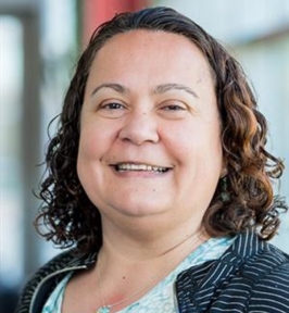 Portrait of Dilek Hakkani-Tür smiling at the camera. She has shoulder-length curly hair and is wearing a patterned top with a dark, striped cardigan. The background is softly blurred, suggesting an indoor setting.