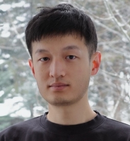 Portrait of Yunze Man looking directly at the camera. He has short dark hair and is wearing a dark crewneck shirt. The background shows softly blurred trees and branches, suggesting an outdoor or window-lit natural setting.