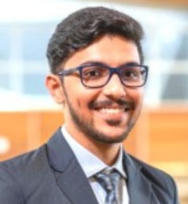 Portrait of Rohan Marwaha smiling indoors. He has short dark hair, a neatly trimmed beard, and is wearing glasses, a dark suit jacket, dress shirt, and tie. The background is softly blurred, suggesting a professional or academic setting.