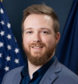 Portrait of Hans Müller Paul smiling at the camera. He has short light brown hair and a full beard, and is wearing a blazer over a dark shirt. A U.S. flag and dark drapery are visible in the background, giving the image a formal, official setting