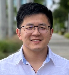 Portrait of Ziqi Pang smiling at the camera. He has short dark hair and is wearing glasses and a light-colored button-down shirt. The background is softly blurred greenery and a walkway, suggesting an outdoor campus or professional setting.