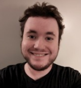 Portrait of Sol Robinson smiling at the camera. He has short, slightly tousled dark hair and a short beard, and is wearing a black T-shirt. The background is a plain, light-colored wall, giving the image a casual, informal headshot appearance.