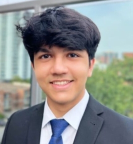 Portrait of Samyak Shrimali smiling at the camera. He has thick dark hair and is wearing a dark suit jacket, white dress shirt, and blue tie. The background shows softly blurred buildings and greenery, suggesting an indoor setting with a city or campus view.