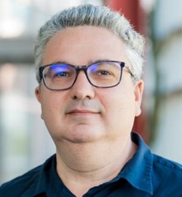Portrait of Gokhan Tür looking at the camera. He has short gray hair and is wearing dark-framed glasses and a dark blue collared shirt. The background is softly blurred, suggesting an indoor professional setting.
