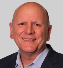 Portrait of John Reid smiling at the camera. He is bald and wearing a dark blazer over a light-colored collared shirt. The background is plain and light gray, giving the image a professional headshot appearance.