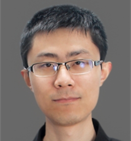 Portrait of Yuxiong Wang looking directly at the camera. He has short dark hair, wears rectangular glasses, and a black collared shirt. The background is plain gray, giving the image a clean, professional headshot appearance.