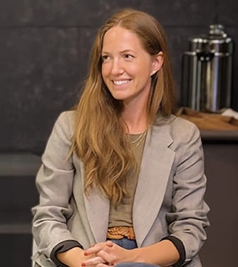 A woman with long brown hair, wearing a gray blazer and a casual top, sits and smiles. The background includes metal containers and a dark wall. (alt text by alttext.ai)