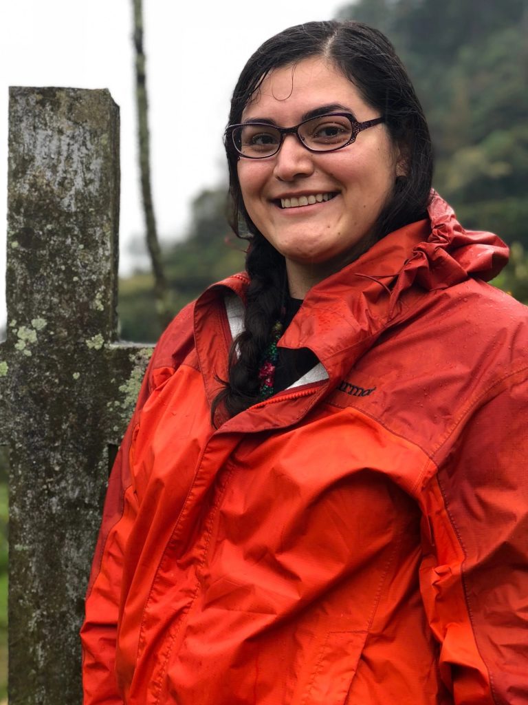 A woman in glasses and a red rain jacket smiles outdoors in front of a weathered stone cross, with a blurred green, misty background. (alt text by alttext.ai)