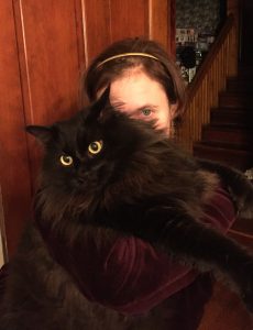 Dana Kinzy peeps out from behind a fluffy black cat with gold eyes.