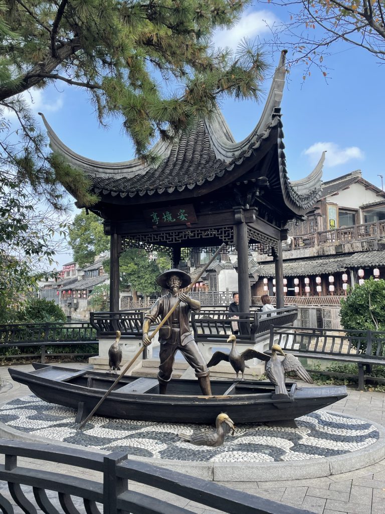 A statue of a man rowing a small boat in front of a pagoda near the river.