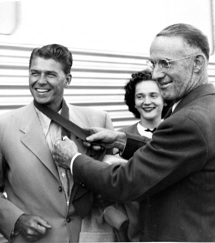 Two men and a woman are standing in front of a train. The older man, known as Mayor Bryon Britt, is tying a tie around the neck of a young Ronald Reagan. A young lady, known as Audrey Hagan, is standing in the background smiling.