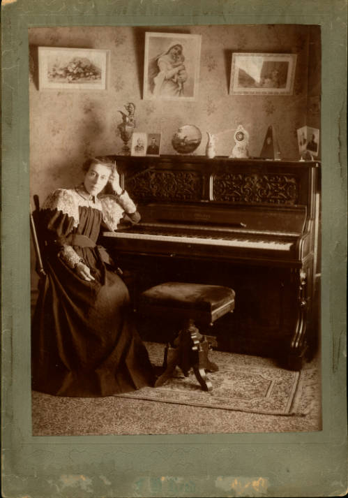 A woman in 19th-century dress poses by an upright piano for a photograph.