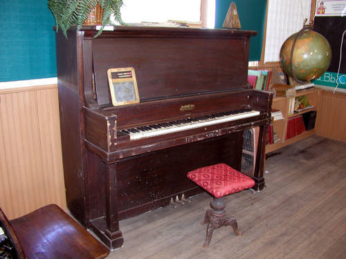 An upright piano stands up against a wall along with other items from a rural one room school house.