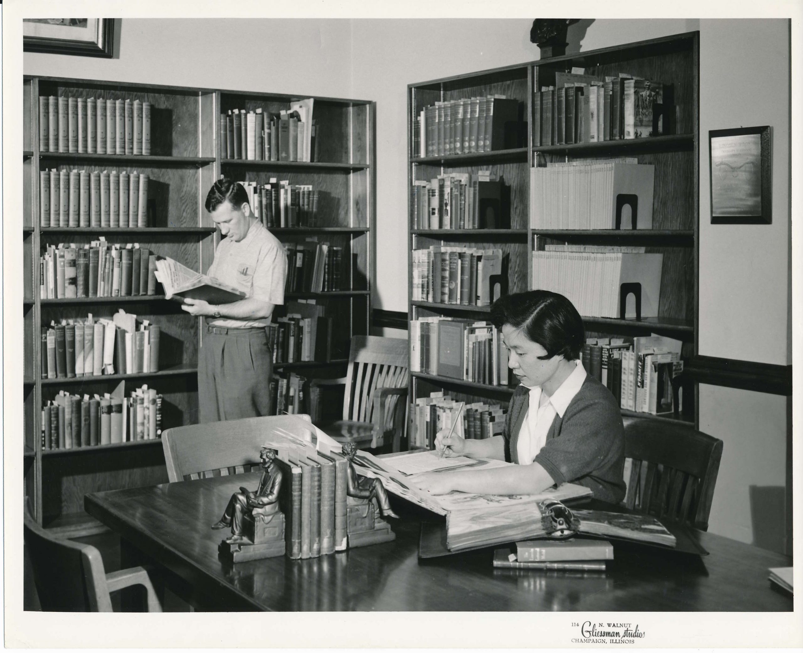 Researchers (one sitting, one standing, both reading books) use the Lincoln Room. Bookshelves line the walls. 