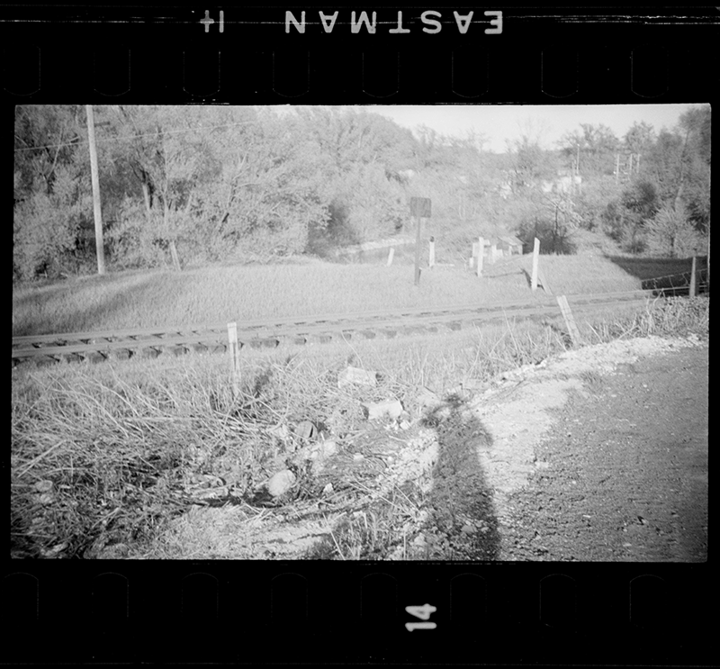 Black and white photo negative of ground with man's shadow. Railroad tracks run through middle of view. Trees in background.