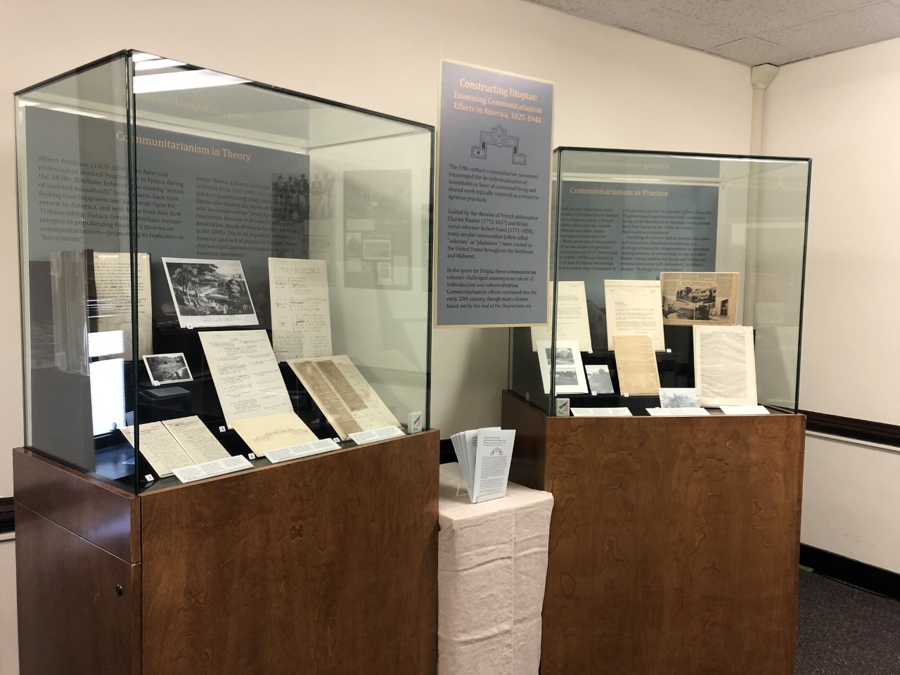 Two glass exhibit cases filled with collection items for exhibition. A hanging poster and table with booklets separate the cases. 