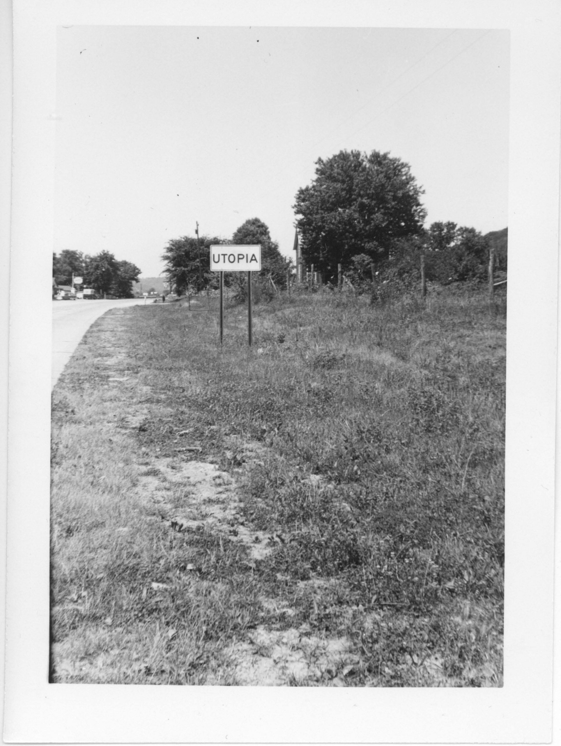 Black and white photograph of a side of a road, with grass, and a single sign reading "UTOPIA." Trees in background.