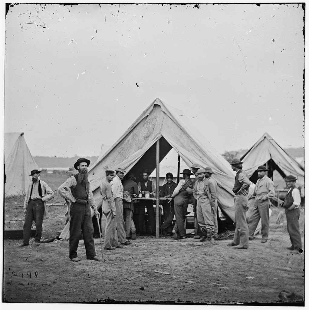 A black and white photograph of men standing outside and in a tent. 