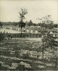 A black and white image of rows of barracks behind a wooden fence in the background. The foreground is a field with a few sparse trees. 