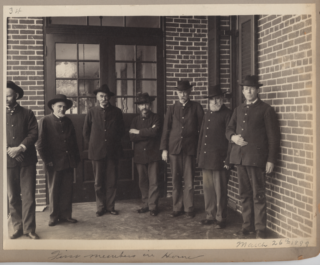 Seven men standing casually in non-military uniforms with brimmed hats. All but one looks to the camera. They are standing in from of a set of doors to a brick building. 