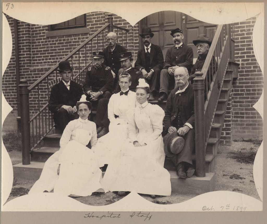 Several men in military uniforms and three women in white nurses' uniforms sit on the steps of a brick building. The photograph is captioned "Hospital Staff" and the date is written in the bottom right corner, "Oct. 7th 1899".