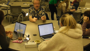 Students with laptops and lab equipment sit at a table talking to their professor.