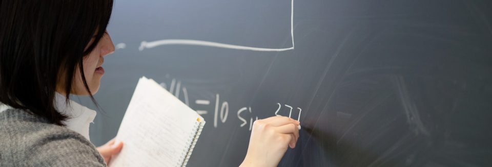 A woman holding a spiral-bound notebook writes an equation on a blackboard.