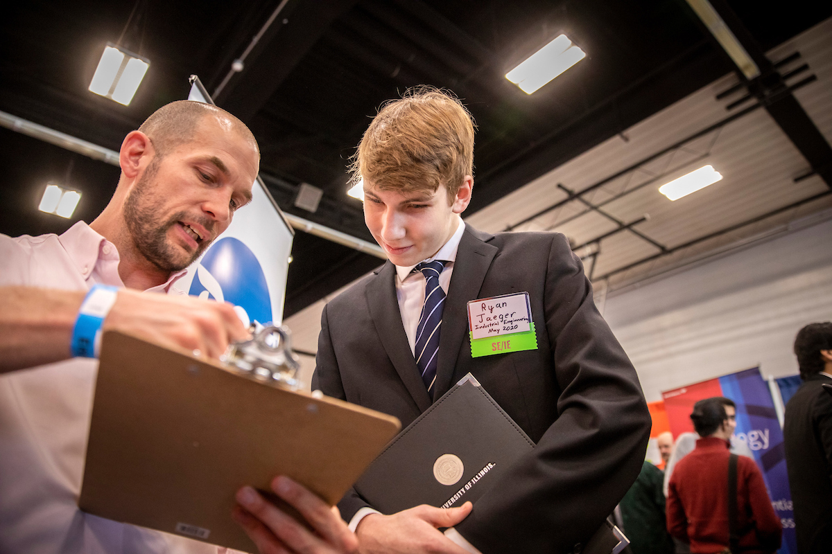 A young man in a business suit pays attention to a clipboard held by a more casually dressed recruiter.