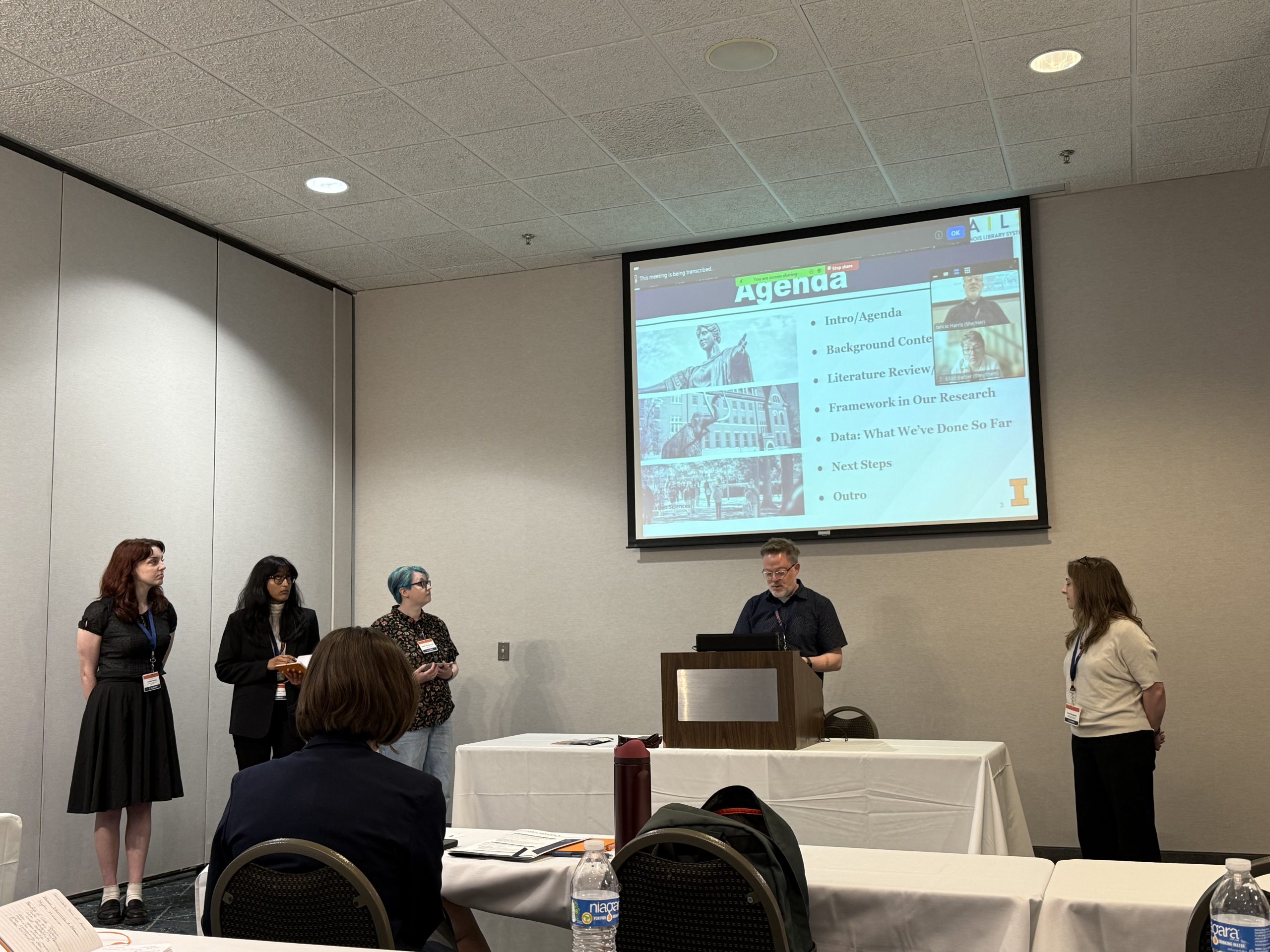 Five people are in a conference room: four standing near a podium and one seated. A person at the podium is presenting a PowerPoint slide titled 
