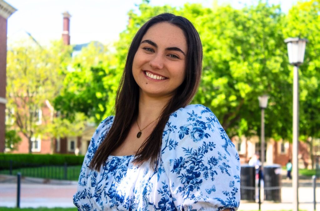 Person wearing a white blouse with blue floral patterns standing outdoors on a sunny day with green trees and campus buildings in the background.