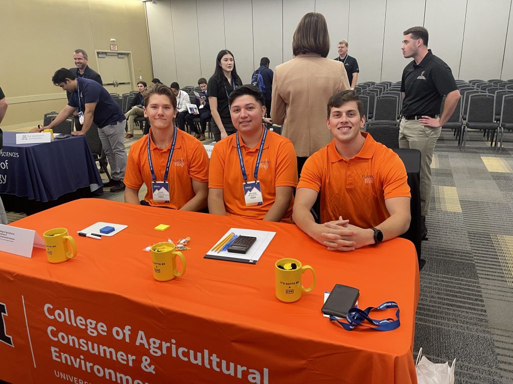 Three individuals wearing matching bright orange polo shirts seated at a table covered with a cloth displaying the text ‘College of Agricultural, Consumer & Environmental Sciences, University of Illinois Urbana-Champaign.’ The table holds yellow mugs, a calculator, papers, and lanyards, with other people and tables visible in the background at an event.