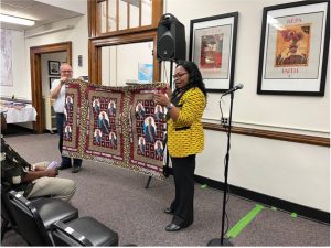 Photo of Émilie Songolo and a man holding up an example of an African textile. The textile is burgundy with a blue pattern.