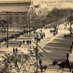 Black and white postcard of long view of people walking in street and on sidewalks across RR tracks. Pullman factory seen in distance