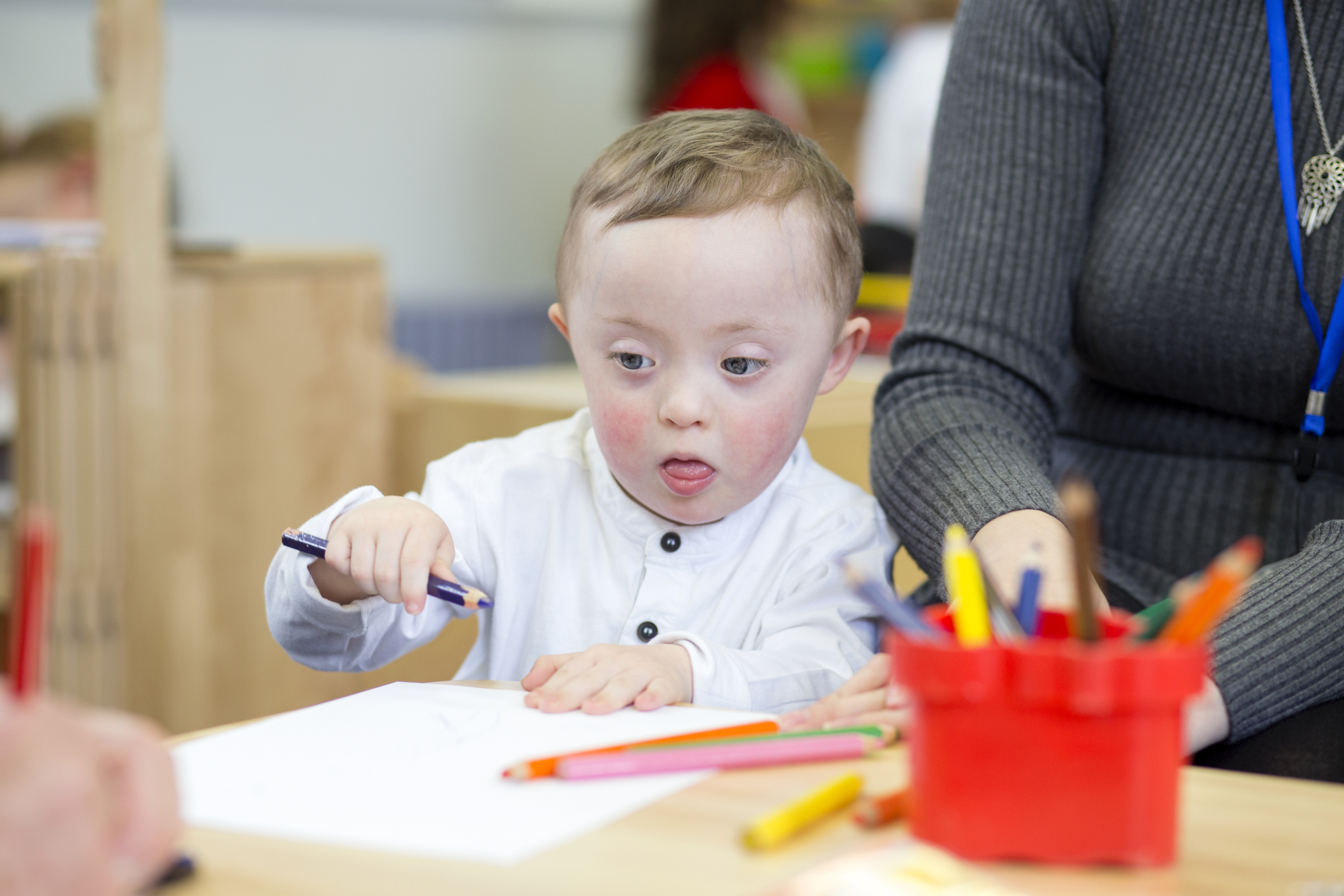 Down Syndrome child doing coloring in at his nursery. His teacher is sitting next to him.