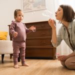 Woman blowing bubbles in the air in the living room while toddler girl looks on.
