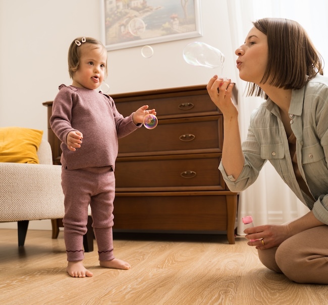 Woman blowing bubbles in the air in the living room while toddler girl looks on.