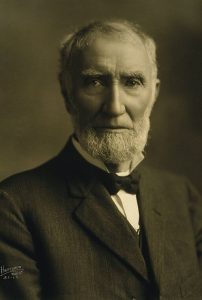 A black and white portrait of an older man with a short white beard, wearing a suit jacket and bow-tie. 