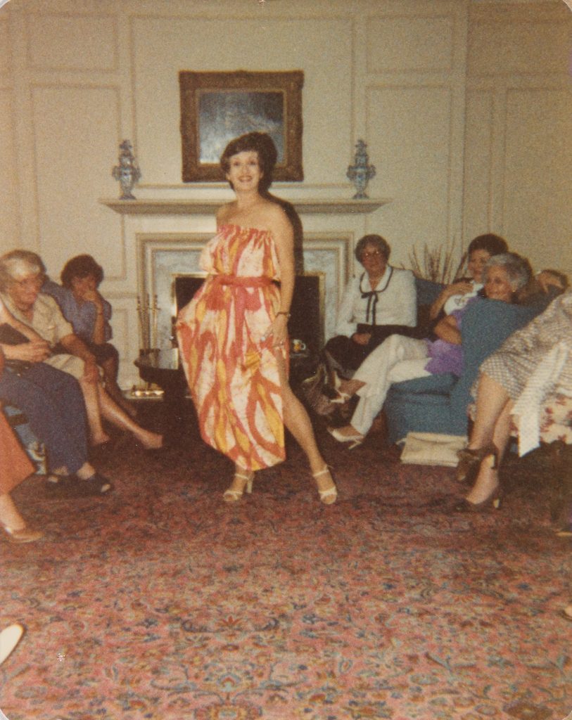 A woman wearing a colorful red, yellow, and white sleeve-less dress poses for a picture. She is surrounded by a number of seated women, who are looking at the woman's dress.