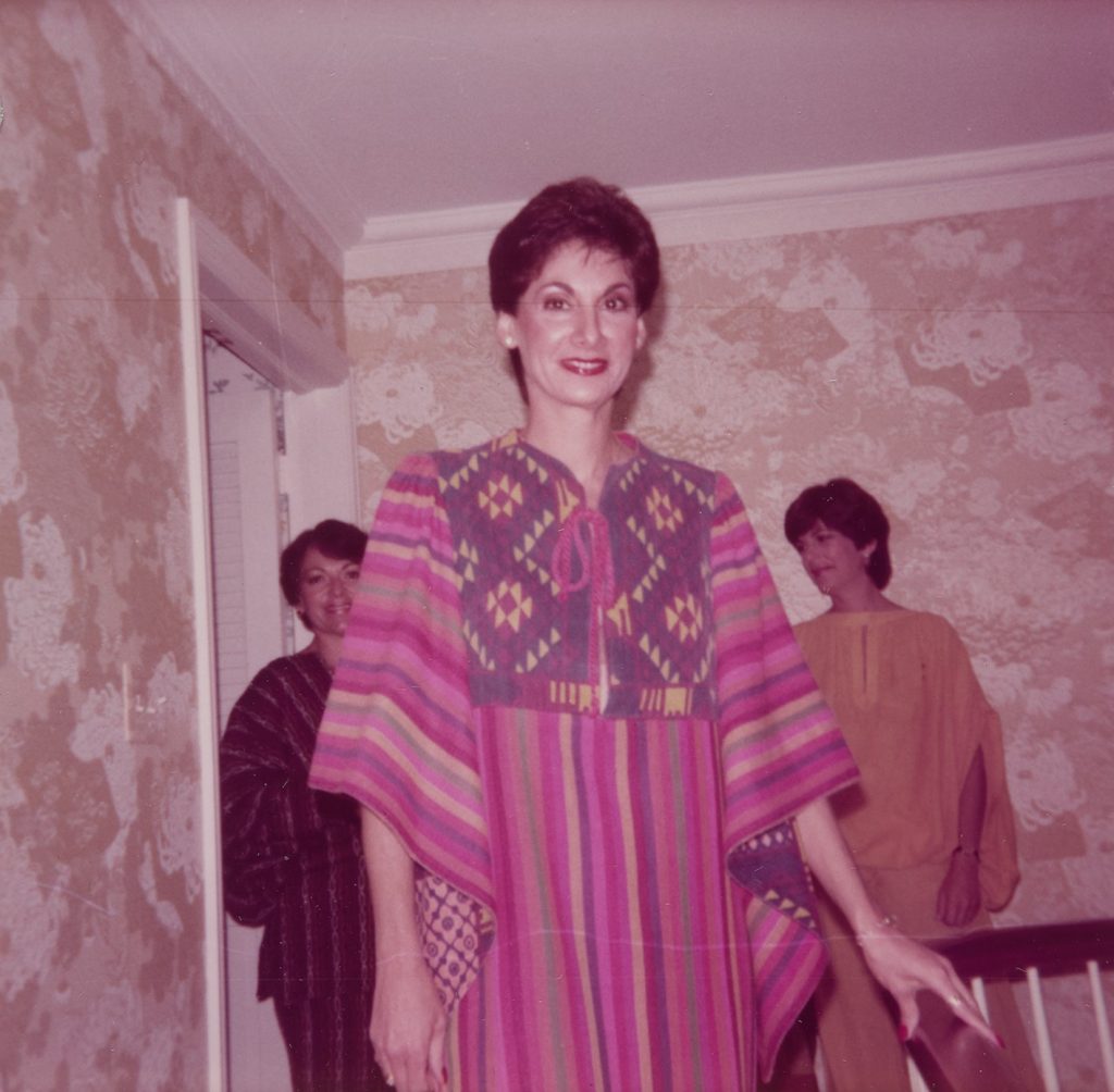 A woman wearing a elaborately patterned, colorful dress poses for a picture. Two woman wearing dresses stand behind her.