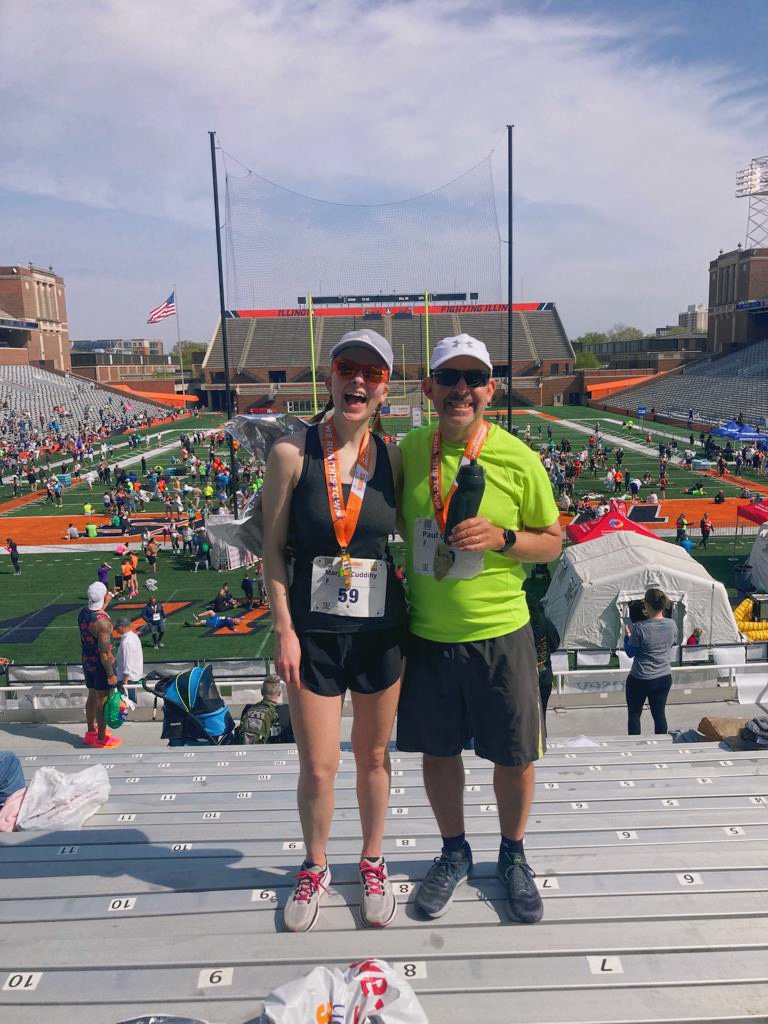 Margot (left) and her dad (right) pose on the stands of Memorial Stadium with the field in the background. They are wearing their half marathon medals.