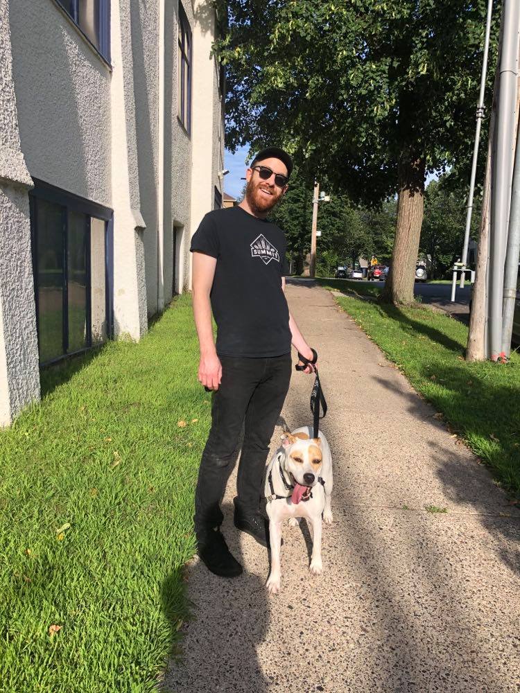 Jonas, a red-haired, bearded man wearing sunglasses and all black, walks a dog.